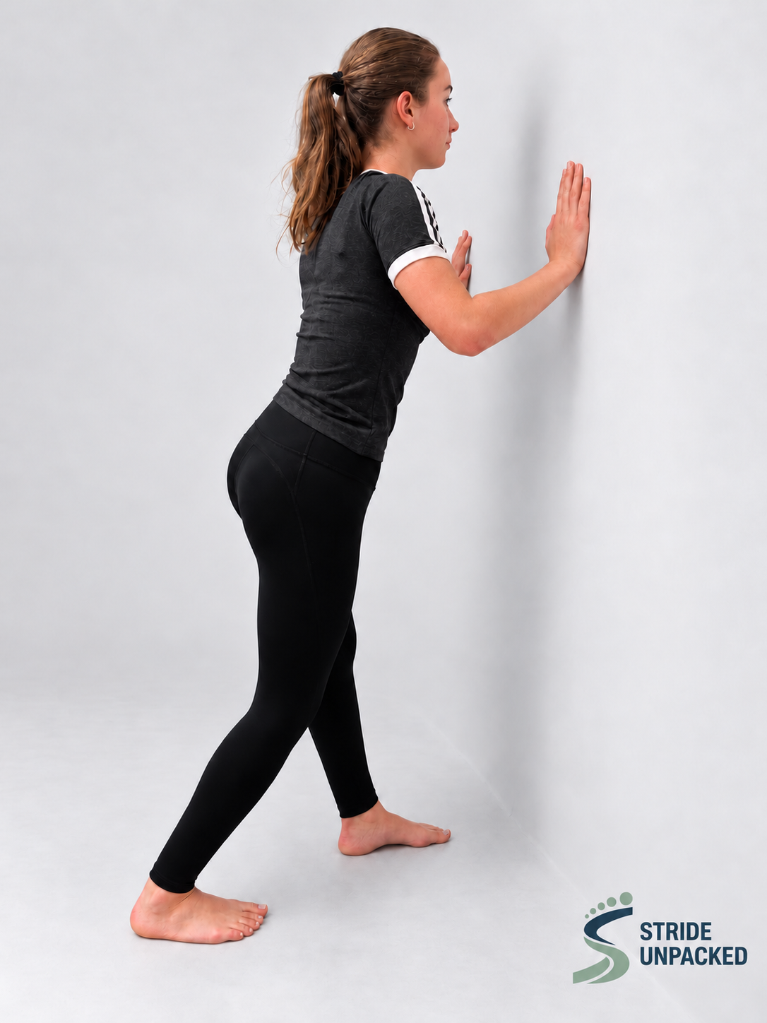 Person demonstrating a wall-supported soleus stretch with the back knee bent and heel on the floor.