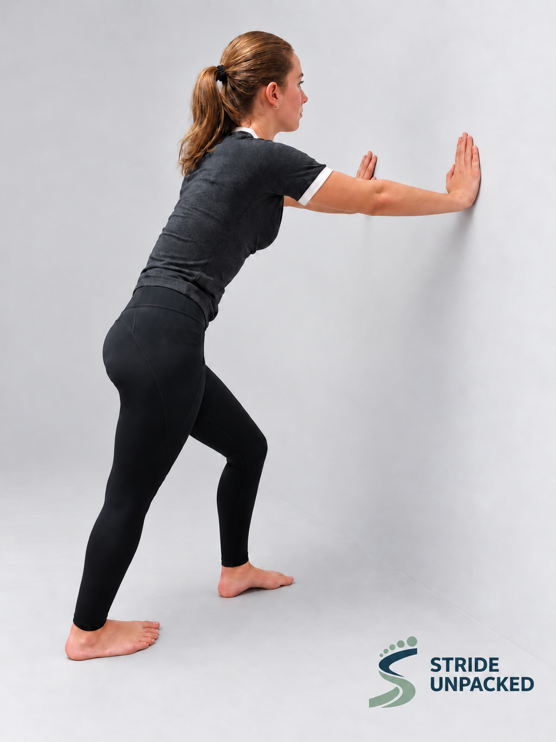 Person demonstrating a wall-supported calf stretch with the front knee bent, back knee straight and back heel on the floor.